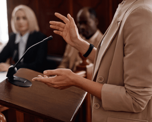 Crop shot of young female witness in beige blazer, standing by tribune with microphone and testifying in front of jury, judge and protection side