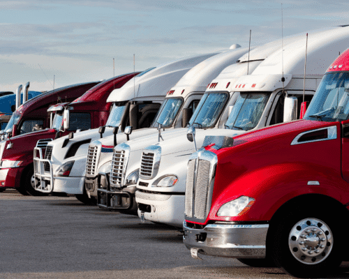 Row of parked semi-trucks in various colors, lined up at a commercial lot under a clear sky