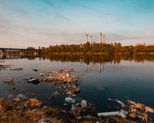 Polluted river with floating trash and industrial smokestacks in the background, symbolizing environmental tort lawyer Columbus GA. The image highlights toxic exposure lawsuit risks, water contamination GA, hazardous waste injury, and air quality claims linked to industrial pollution.
