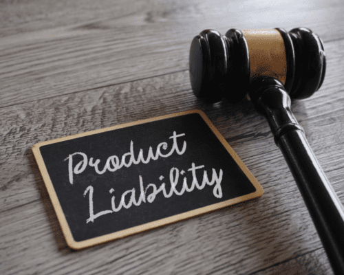 A wooden judge’s gavel lies beside a small chalkboard with the handwritten words “Product Liability.” The image visually represents legal accountability for defective or dangerous products, emphasizing manufacturer responsibility and consumer safety.