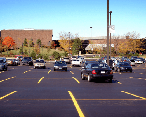 A large parking lot with several parked cars on a clear day, bordered by trees with autumn foliage and a brick building in the background. The image represents public and commercial property where parking lot injuries or premises liability incidents could occur.
