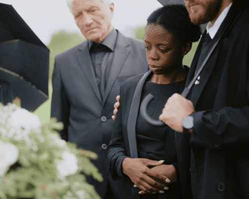 A group of mourners dressed in black stand together under umbrellas at a funeral, with one woman being comforted by others. The image represents the emotional weight of wrongful death cases and the pursuit of justice for families who have lost loved ones due to negligence.
