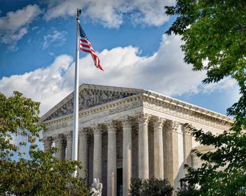 The U.S. Supreme Court building, viewed from below with the American flag waving in the foreground and green trees framing the scene. The image represents justice and federal authority, supporting a legal discussion about a Supreme Court case involving private military contractor liability for personal injury claims.