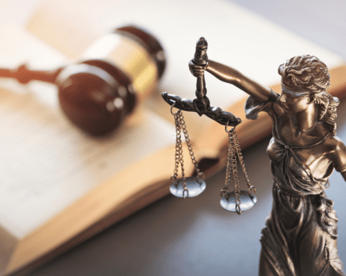A bronze statue of Lady Justice holding balanced scales stands beside an open law book and a judge’s gavel. The image symbolizes fairness, truth, and accountability in legal proceedings, representing whistleblower lawsuits that expose healthcare fraud under the False Claims Act.