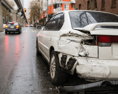 Damaged white sedan with severe rear-end impact sits on a wet city street at a red light, with visible debris on the road and other vehicles driving away in the background.