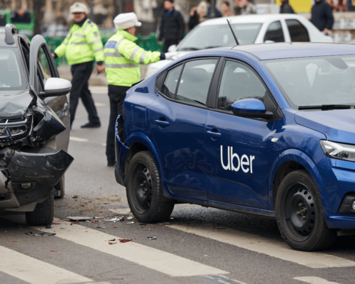 Blue Uber vehicle involved in a rear-end collision at an intersection, with visible front-end damage to another car, debris on the roadway, and police officers directing traffic in the background.