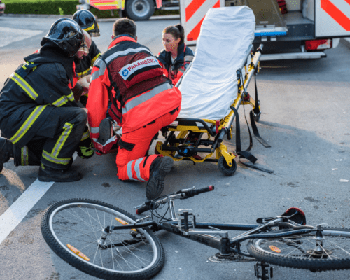 Paramedics and firefighters assist an injured cyclist at the scene of a roadway crash while a damaged bicycle lies nearby.