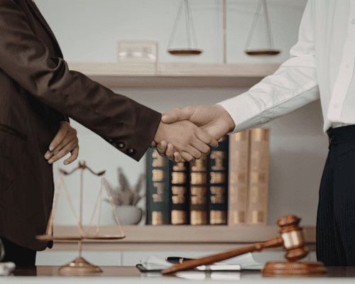 Two professionals shake hands across a desk with legal books, scales of justice, and a gavel in the background, symbolizing legal representation and whistleblower protection.